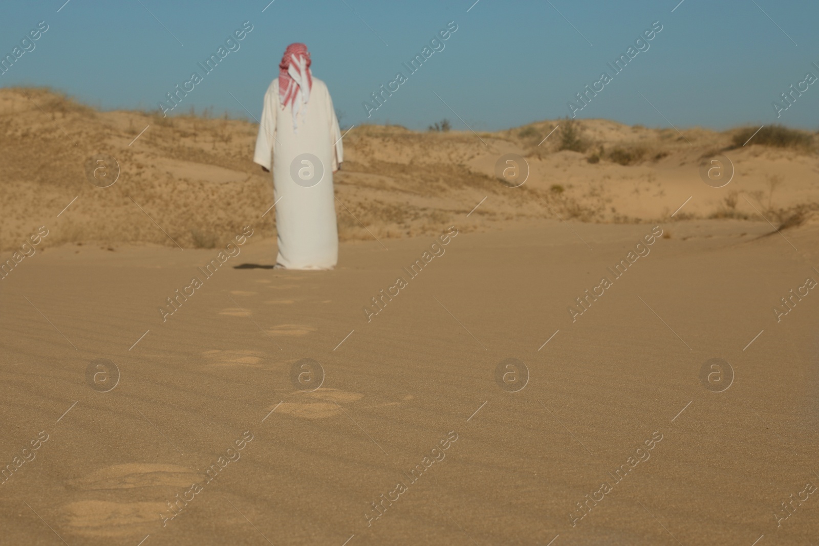 Photo of Man in arabic clothes walking through desert on sunny day, back view