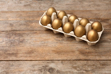 Carton with golden eggs on wooden table, space for text