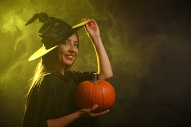 Photo of Young woman wearing witch costume with pumpkin in smoke cloud on dark background, space for text. Halloween party