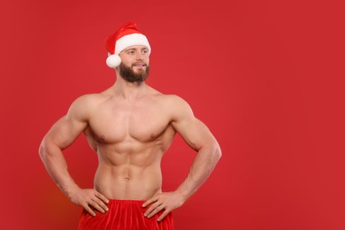 Attractive young man with muscular body in Santa hat on red background, space for text