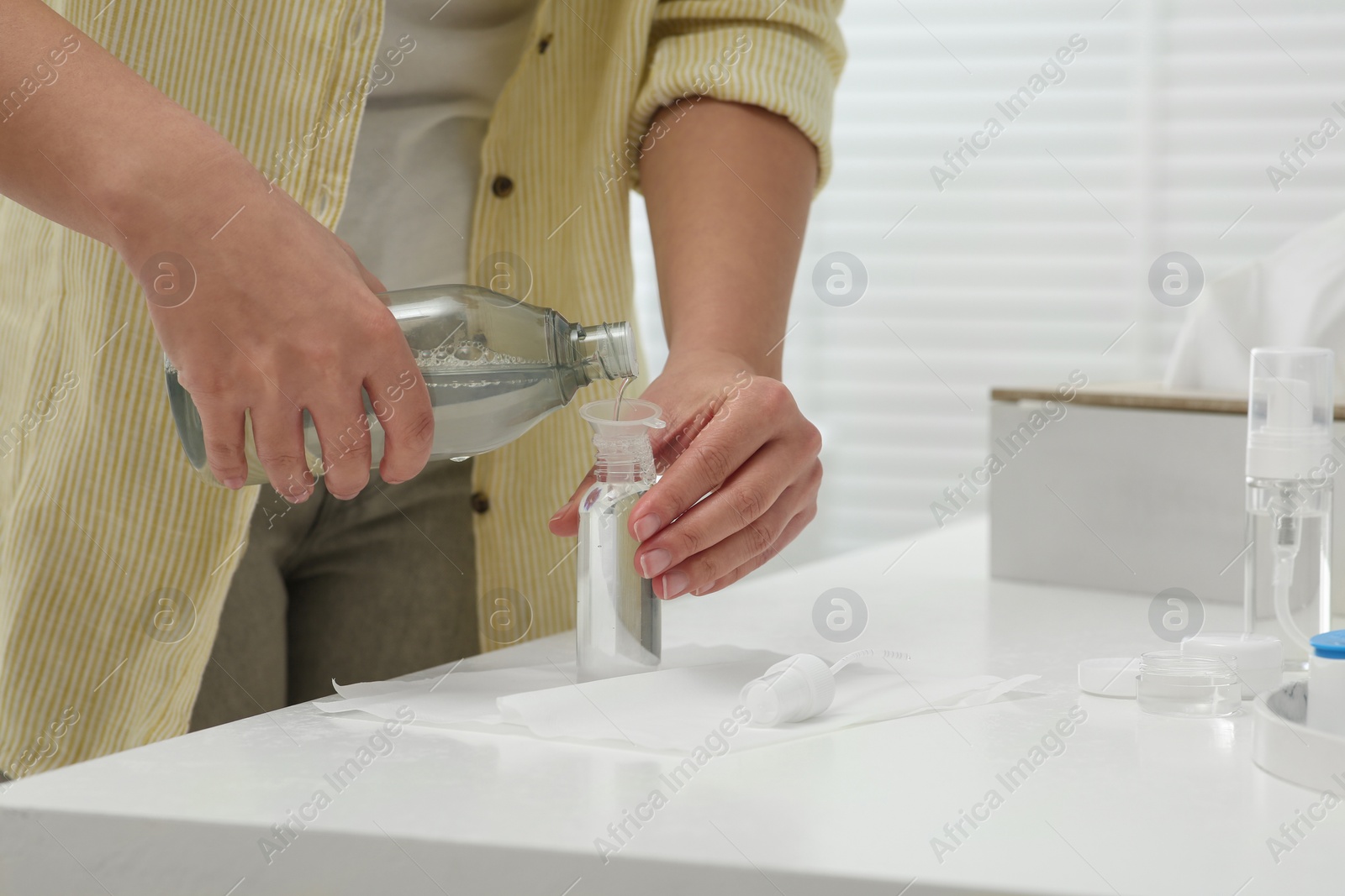 Photo of Woman pouring cosmetic product into plastic bottle at white countertop in bathroom, closeup. Bath accessories