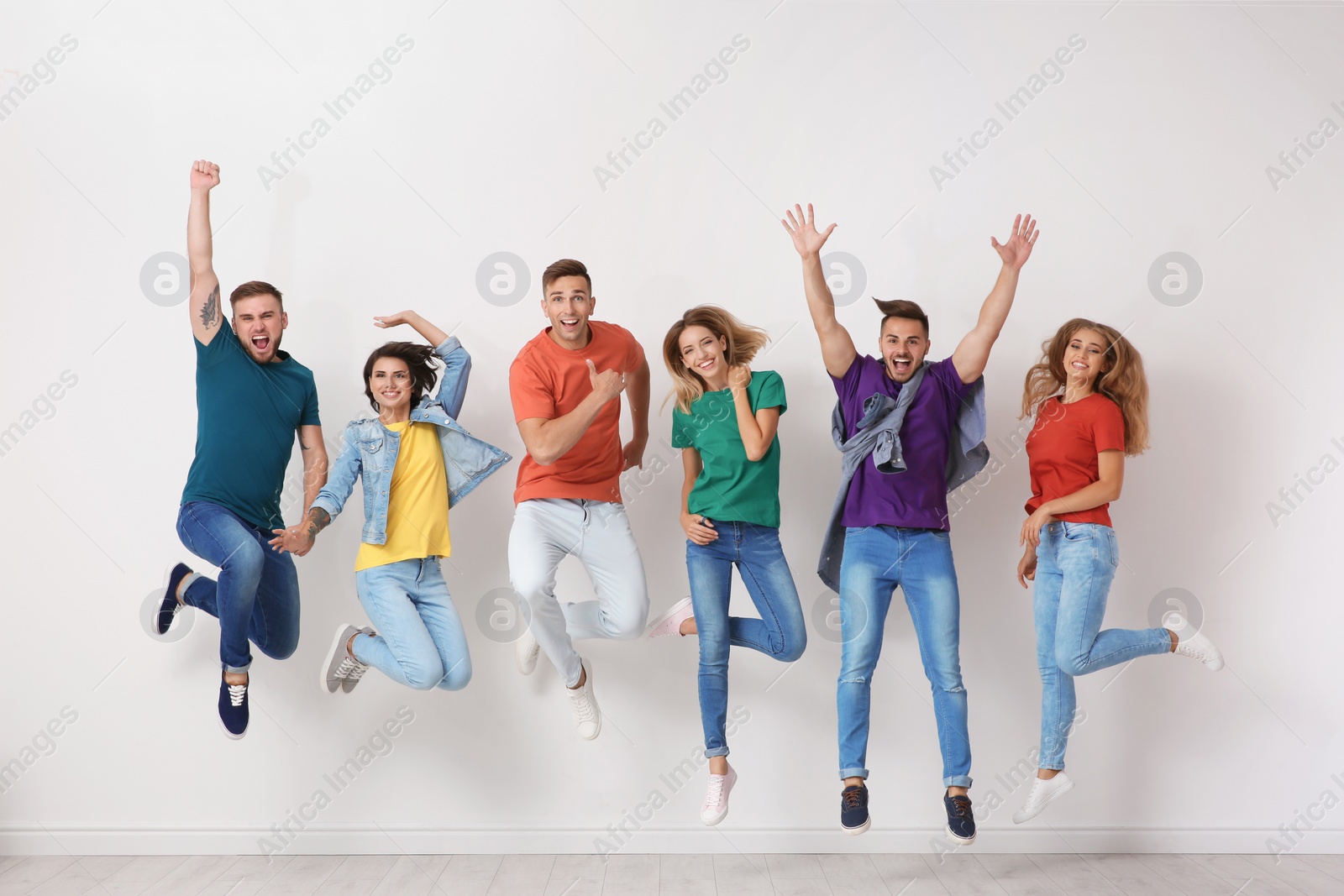 Photo of Group of young people in jeans and colorful t-shirts jumping near light wall