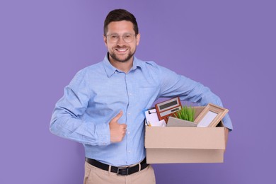 Happy unemployed man with box of personal office belongings showing thumb up on purple background