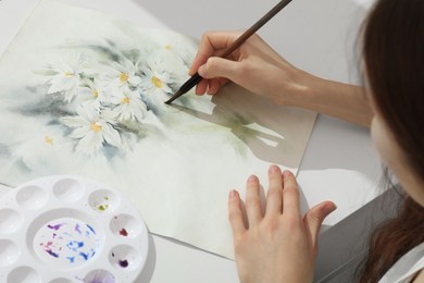 Photo of Woman painting flowers with watercolor at white table, above view. Creative artwork