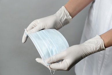 Doctor in medical gloves holding protective mask on light grey background, closeup