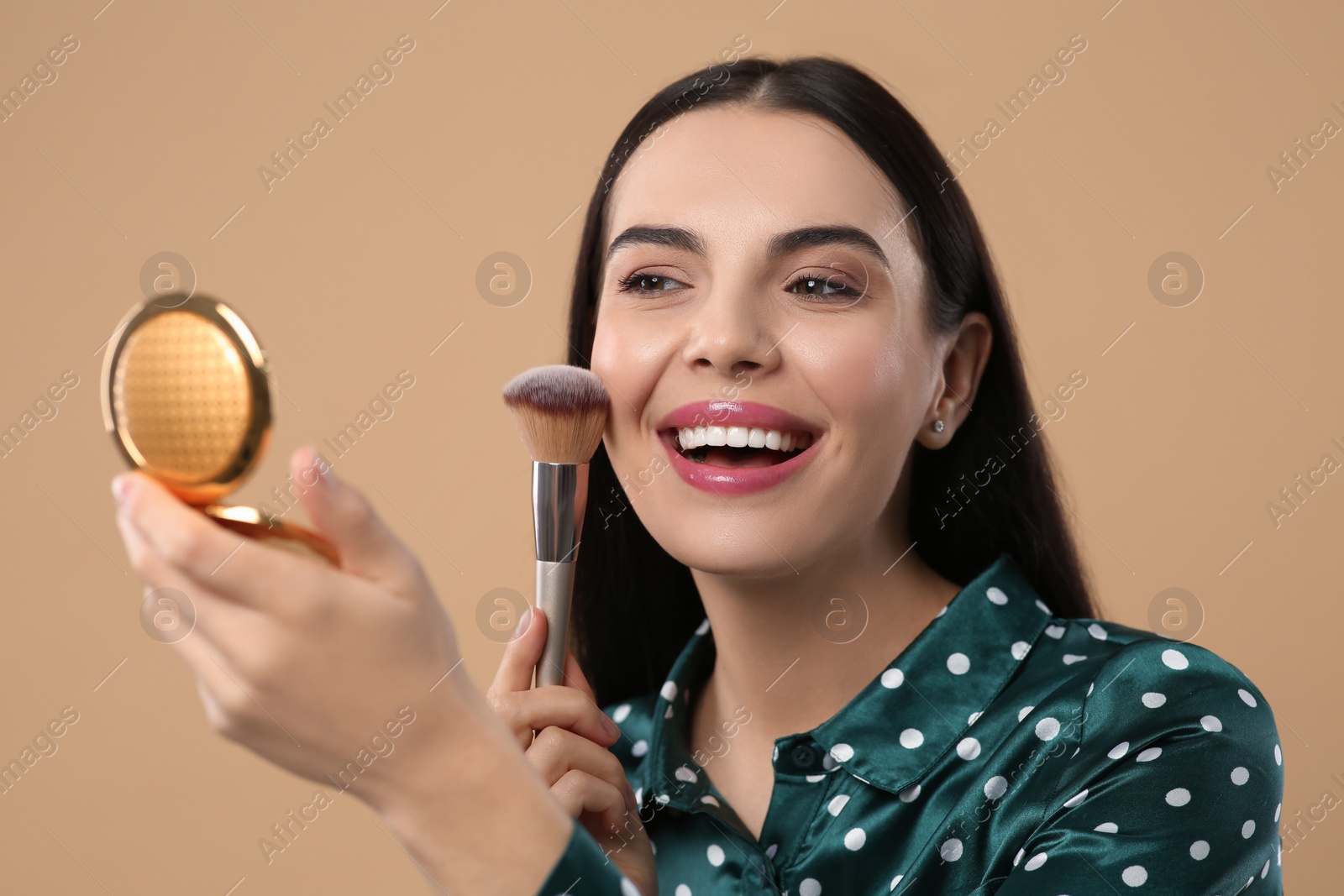 Photo of Happy woman with cosmetic pocket mirror applying makeup on light brown background