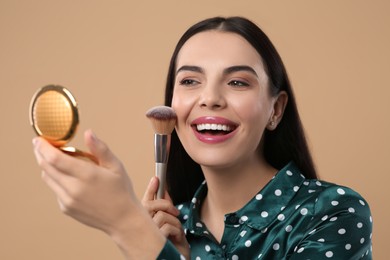 Happy woman with cosmetic pocket mirror applying makeup on light brown background