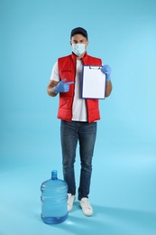 Photo of Courier in face mask with clipboard and bottle of cooler water on light blue background. Delivery during coronavirus quarantine