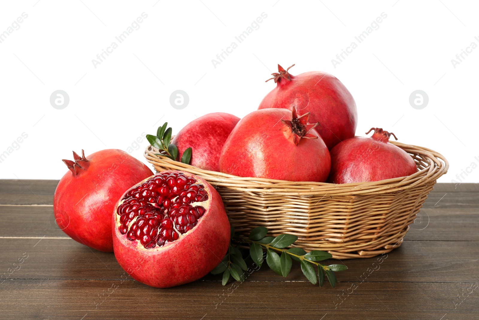 Photo of Fresh pomegranates in wicker basket and green leaves on wooden table against white background