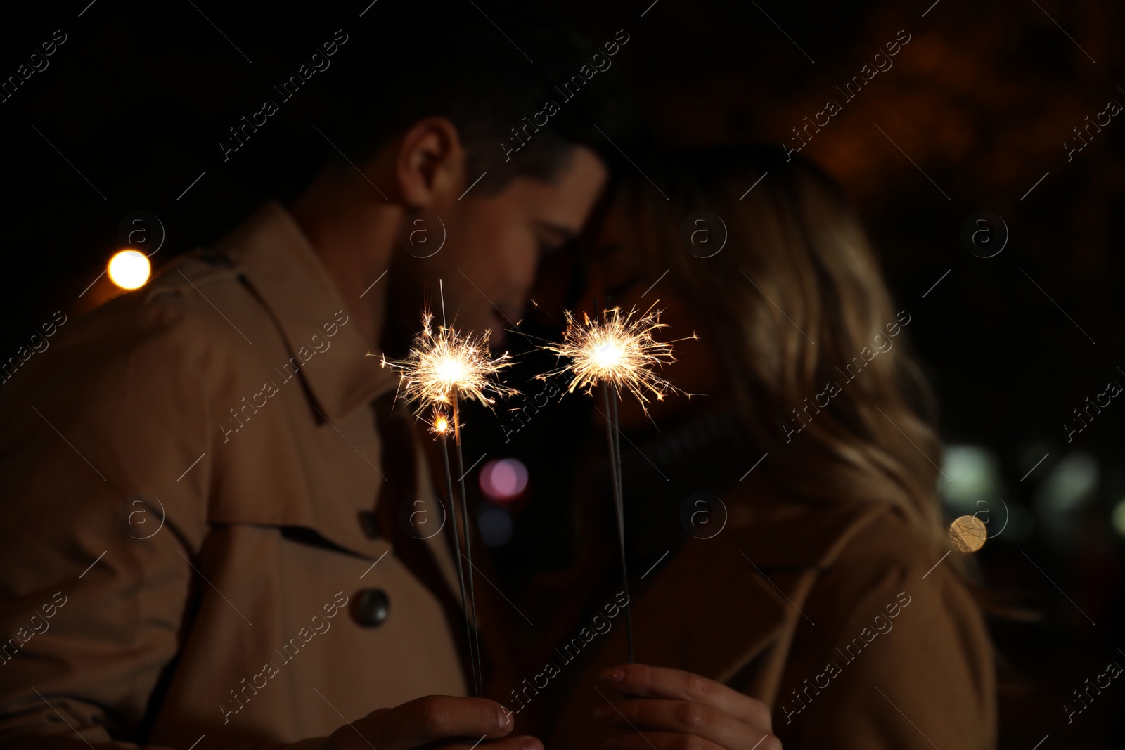 Photo of Couple in warm clothes holding burning sparklers at night, focus on fireworks