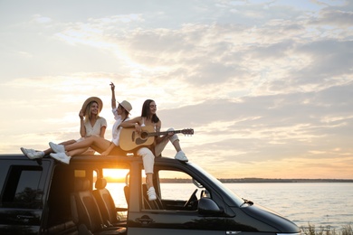 Photo of Happy friends with guitar sitting on car roof outdoors at sunset. Summer trip
