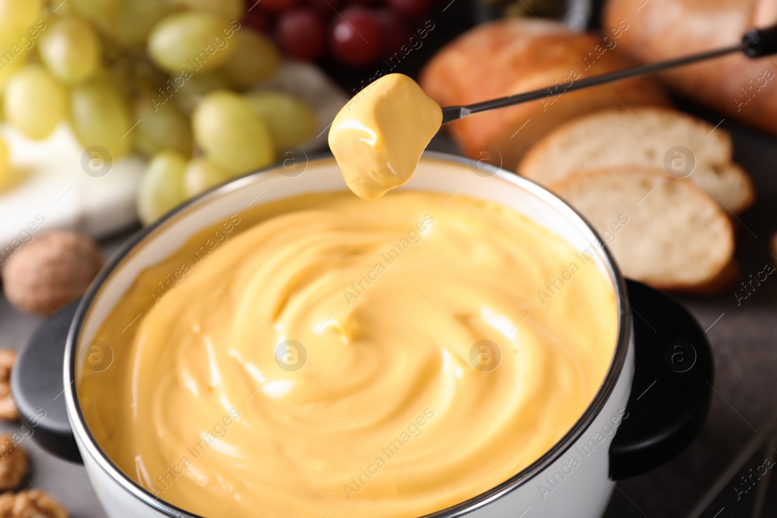 Photo of Piece of bread over pot with delicious cheese fondue on table