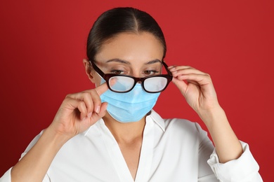 Photo of Woman wiping foggy glasses caused by wearing medical mask on red background