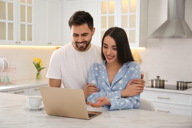 Photo of Happy couple in pajamas with laptop at kitchen table