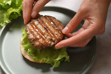 Photo of Woman making tasty hamburger with fried patty, lettuce and bun at table, closeup