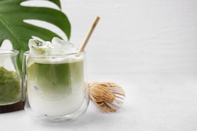 Photo of Glass of tasty iced matcha latte, leaf and bamboo whisk on white table, closeup. Space for text
