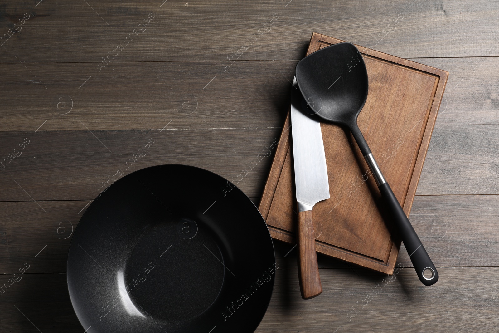 Photo of Black metal wok, knife, board and spatula on wooden table, top view