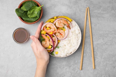 Woman holding delicious poke bowl with shrimps, rice and vegetables at light grey table, top view
