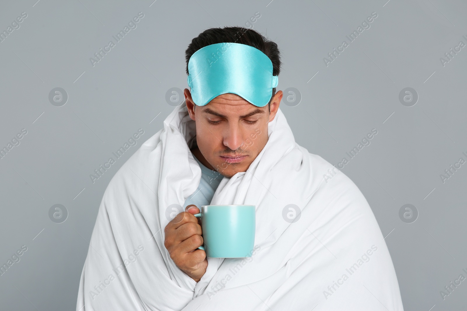 Photo of Man in sleeping mask wrapped with blanket holding cup on grey background