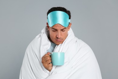 Man in sleeping mask wrapped with blanket holding cup on grey background