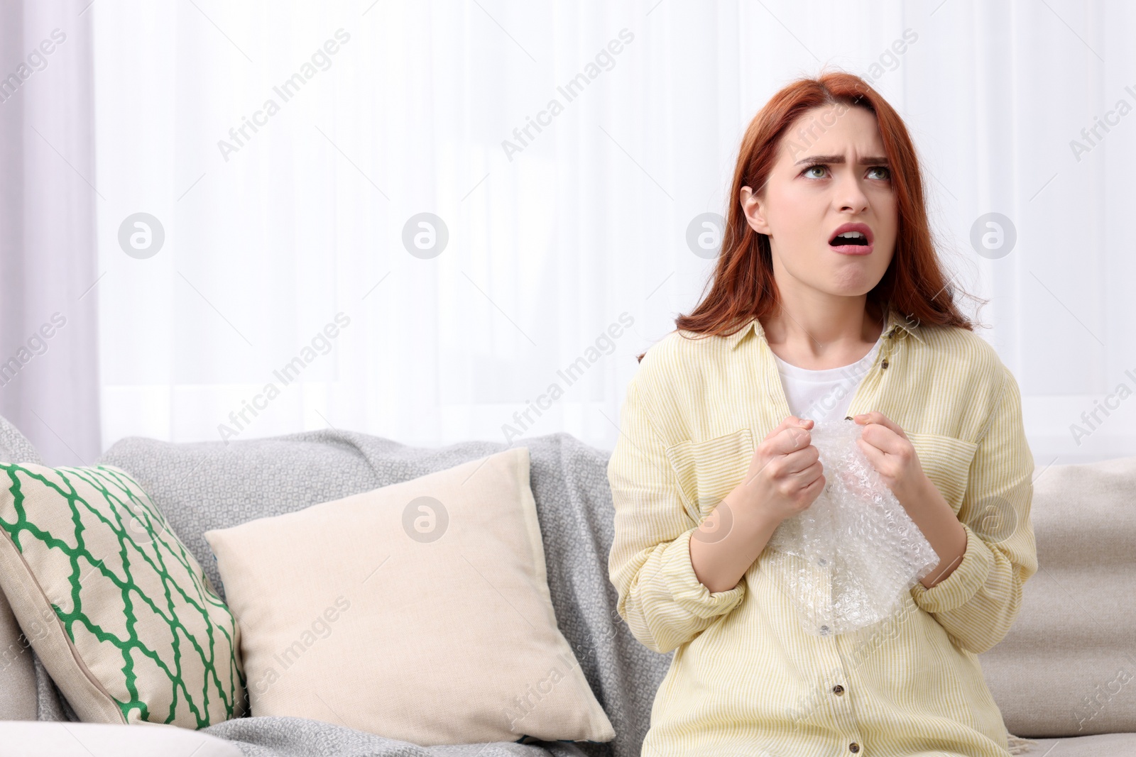 Photo of Angry woman popping bubble wrap on sofa at home, space for text. Stress relief