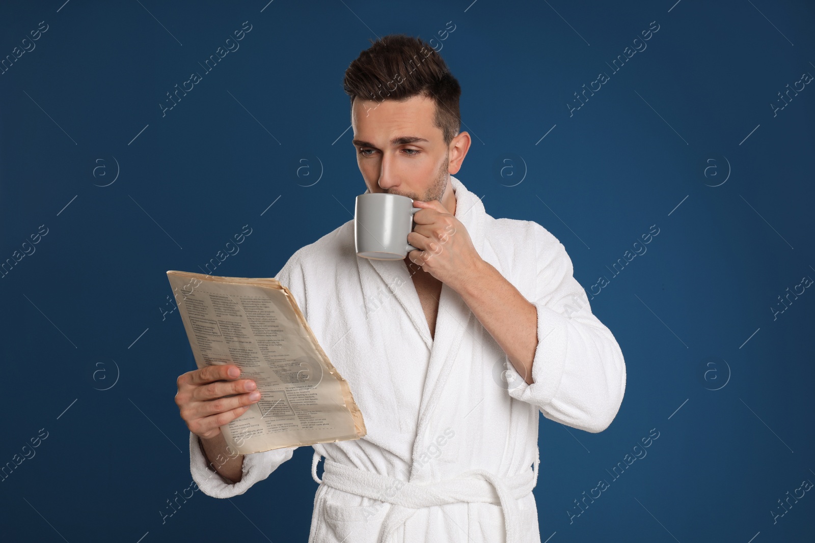 Photo of Young man in bathrobe with cup of coffee reading newspaper on blue background