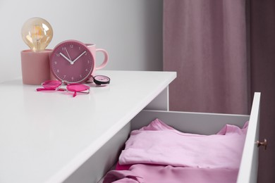 White chest of drawers with pink clothes, clock, sunglasses and decor indoors