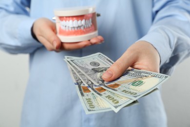 Photo of Woman holding educational dental typodont model and dollar banknotes on light background, closeup. Expensive treatment