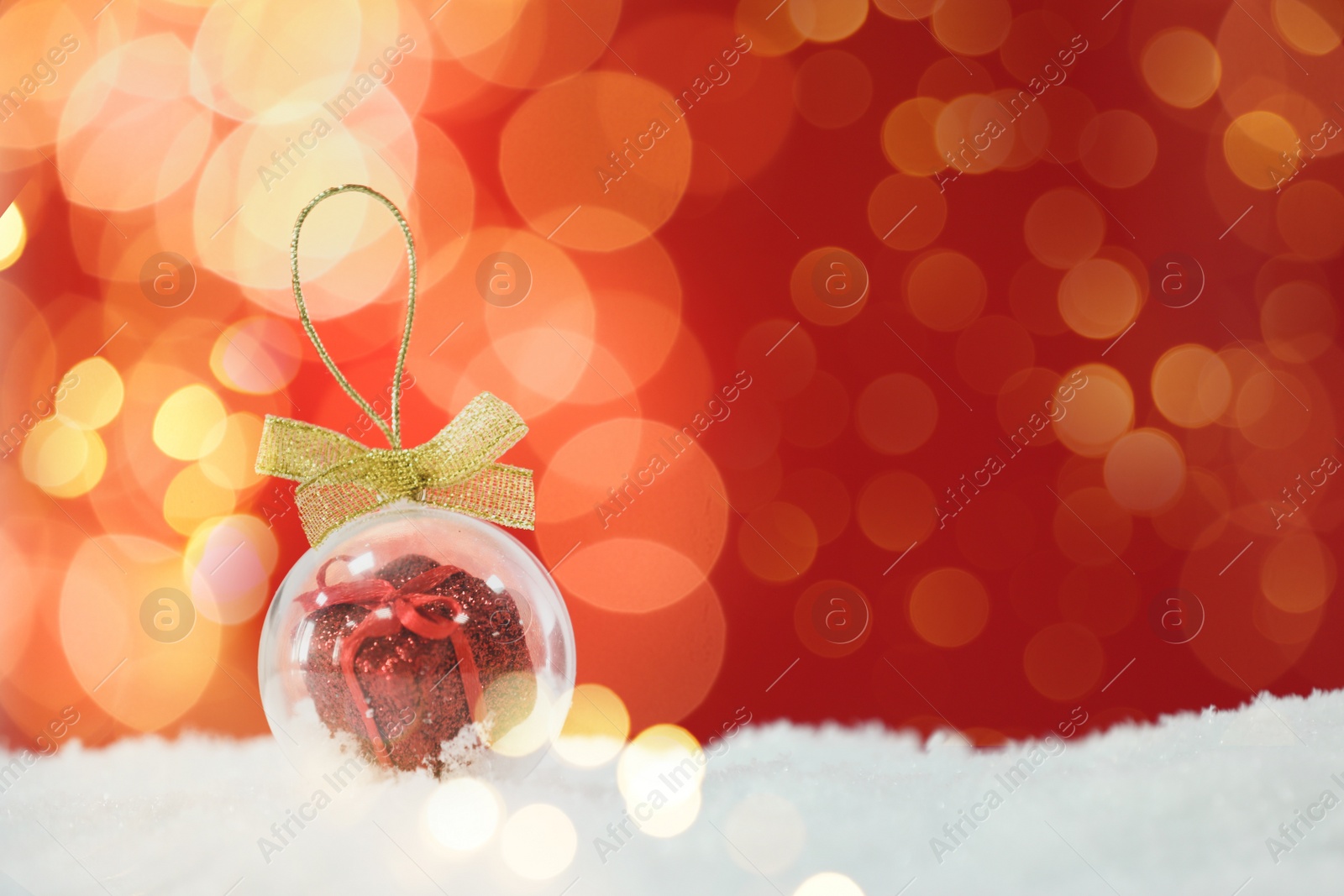 Image of Beautiful transparent Christmas ornament with small gift box on snow against red background, bokeh effect. Space for text