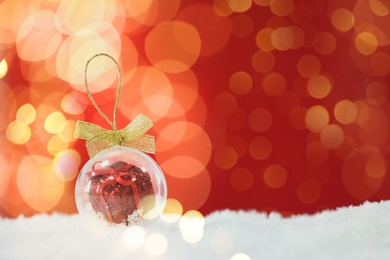 Image of Beautiful transparent Christmas ornament with small gift box on snow against red background, bokeh effect. Space for text