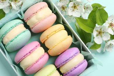 Photo of Many delicious colorful macarons in box and flowers on light blue background, flat lay