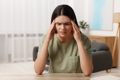 Photo of Sad woman suffering from headache at wooden table indoors