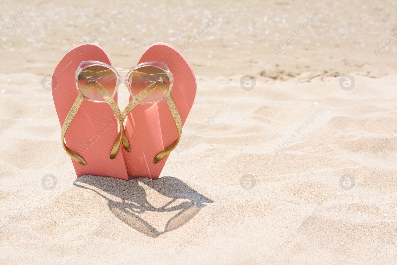 Photo of Stylish pink flip flops with sunglasses in sand on sunny day, space for text