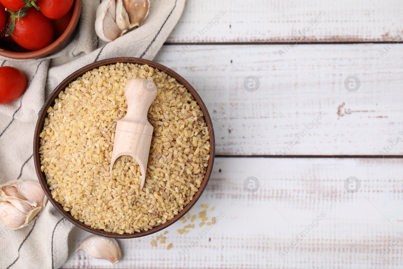 Photo of Bowl of raw bulgur with scoop, tomatoes and garlic on white wooden table, flat lay. Space for text