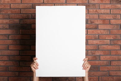 Woman holding white blank poster near red brick wall, closeup. Mockup for design