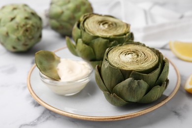 Photo of Delicious cooked artichokes with tasty sauce served on white marble table, closeup
