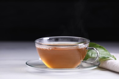 Photo of Aromatic hot tea in glass cup and leaf on white table against black background