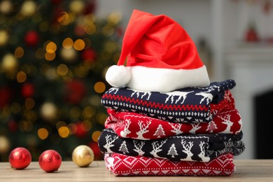 Photo of Stack of different Christmas sweaters, Santa Claus hat and decorative balls on table against blurred lights, space for text