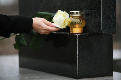 Woman holding white rose near black granite tombstone with candle outdoors, closeup. Funeral ceremony