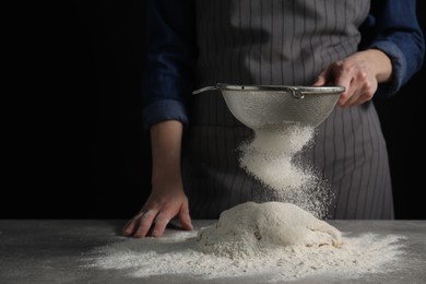 Photo of Making bread. Woman sifting flour over dough at table on dark background, closeup