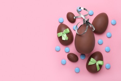 Photo of Delicious chocolate eggs with bows and candies on pink background, flat lay. Space for text