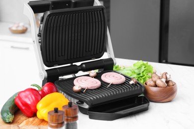 Photo of Electric grill with homemade sausages, mushrooms, and vegetables on white table indoors