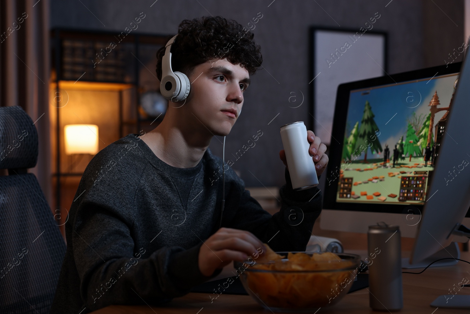 Photo of Young man with energy drink and headphones playing video game at wooden desk indoors