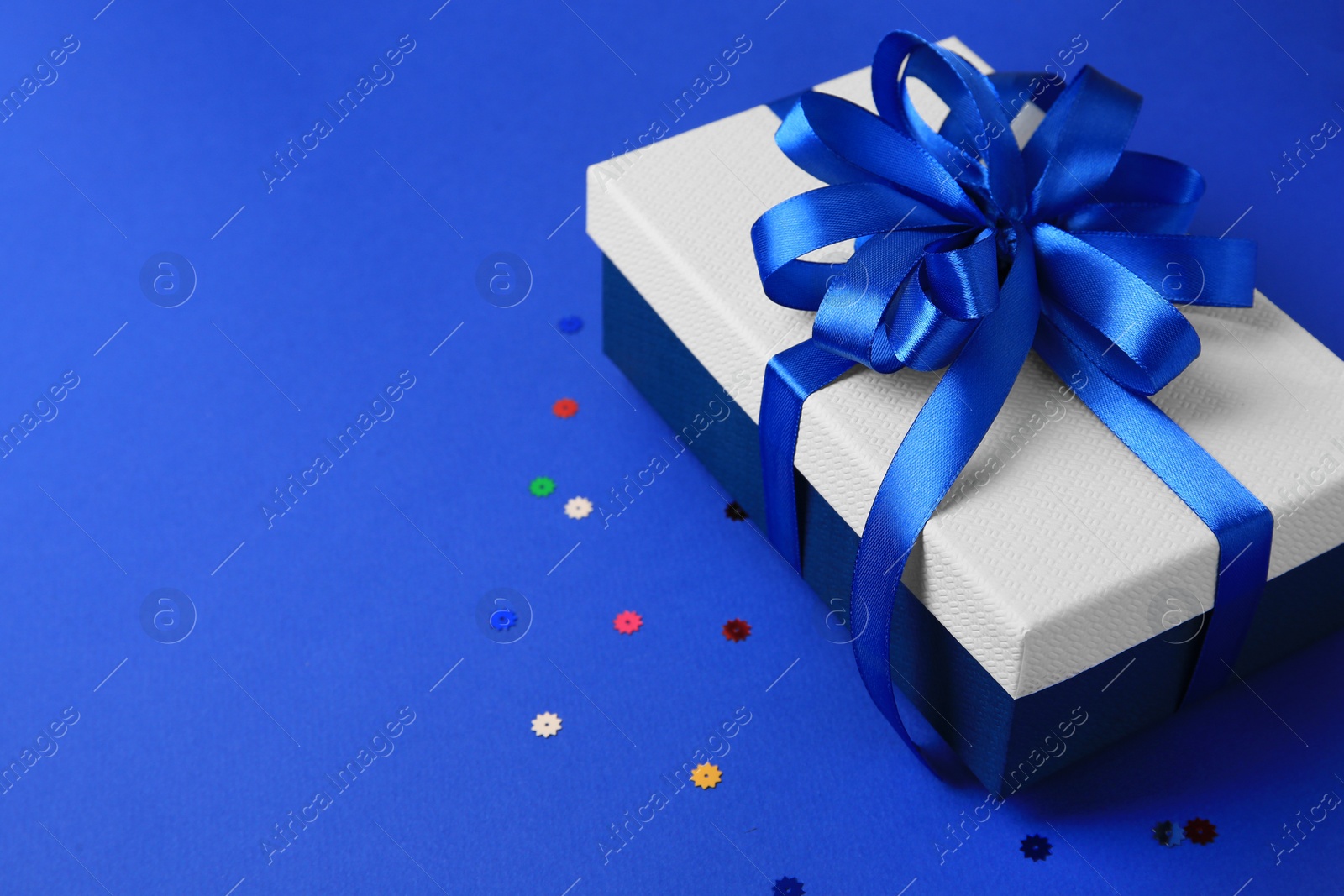 Photo of Beautiful gift box with bow and confetti on blue background. Space for text