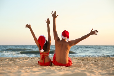 Photo of Lovely couple with Santa hats having fun together on beach, back view. Christmas vacation