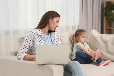 Photo of Woman working remotely at home. Busy mother watching over her daughter on sofa in living room