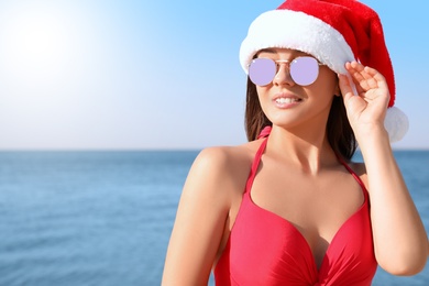 Beautiful young woman in Santa hat and bikini on beach, space for text. Christmas vacation