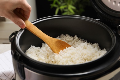 Photo of Woman taking tasty rice with spoon from cooker in kitchen, closeup