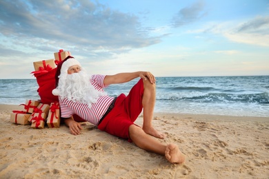 Photo of Santa Claus with bag of presents relaxing on beach, space for text. Christmas vacation