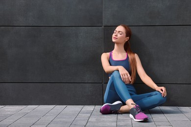 Beautiful woman in stylish gym clothes sitting near dark grey wall on street, space for text